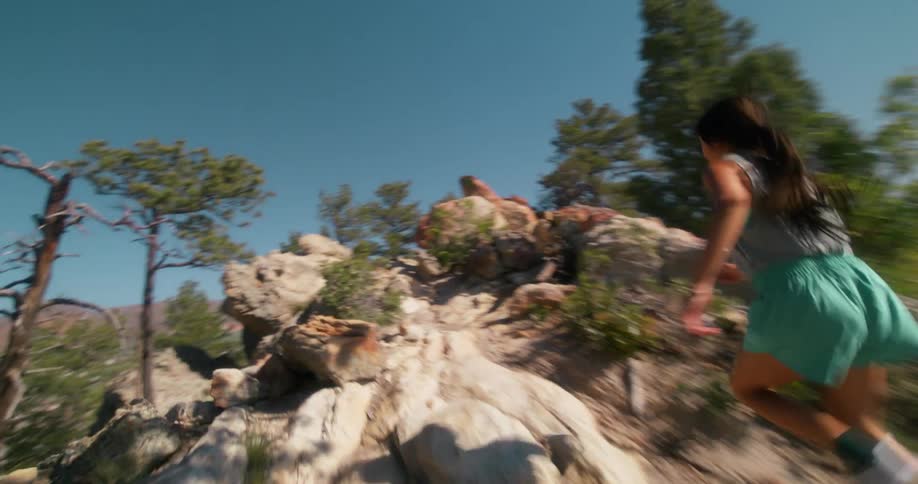 A woman runs along a rocky trail bordered by dry shrubs and trees under a clear blue sky
