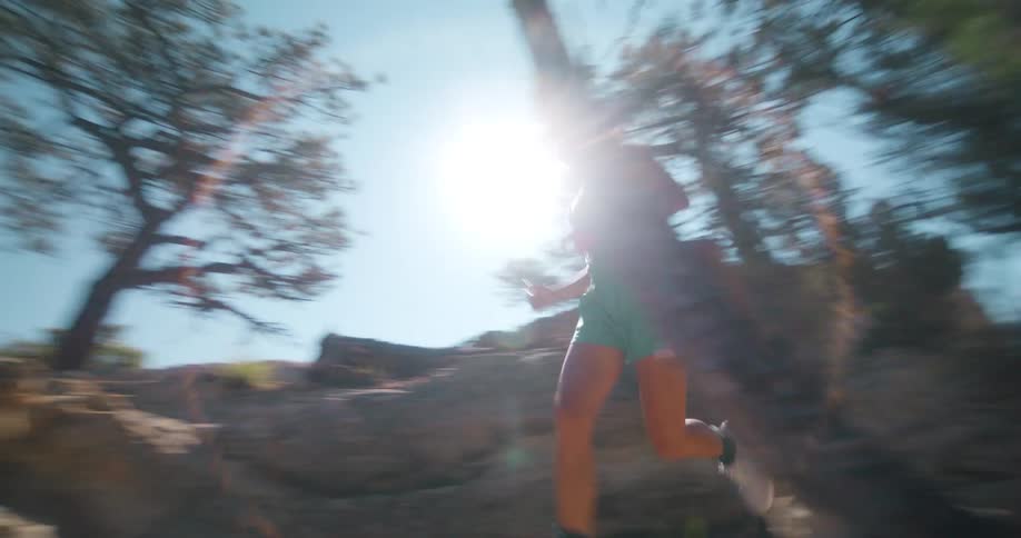 A woman runs on a rocky terrain at midday surrounded by trees