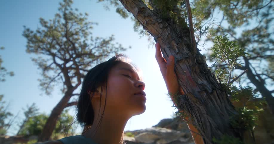 A woman with closed eyes and a peaceful expression leans against a tree trunk with sunlight illuminating her face and green trees in the background