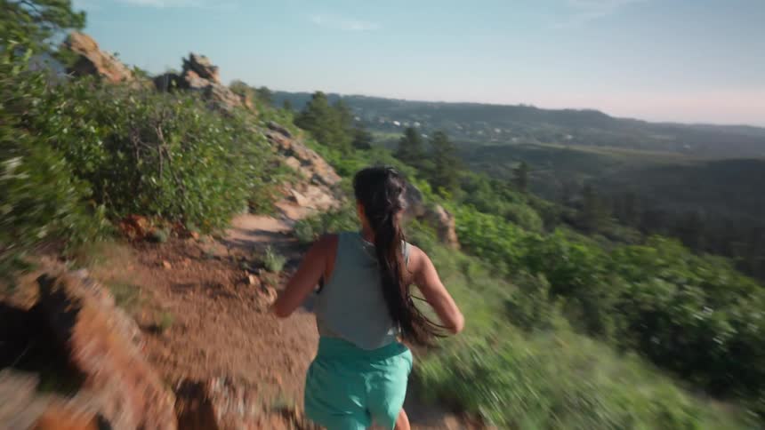 A woman with long hair runs along a rocky trail bordered by dense foliage overlooking a layered vista of forested hills under a clear sky