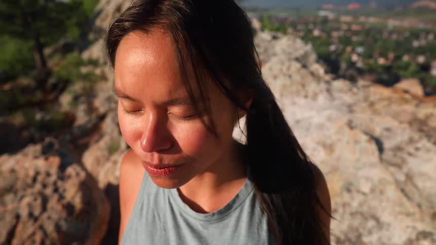A girl is standing near rocks with her eyes closed and breathing deeply