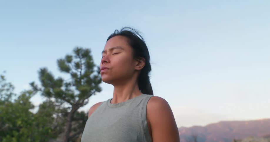 A woman stands outdoors with her eyes closed calmly breathing in the fresh air as the sun rises over the mountains behind her