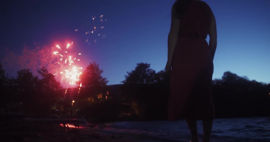 A woman stands on the beach watching a fireworks display at night