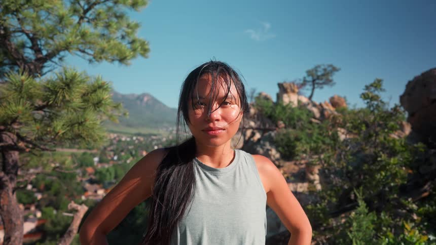 Asian woman who appears to have just finished a hike she has black long hair in a ponytial and her bangs are swaty she is heavily breathing with a picture perfect view pf trees and houses and a mountain in the far background