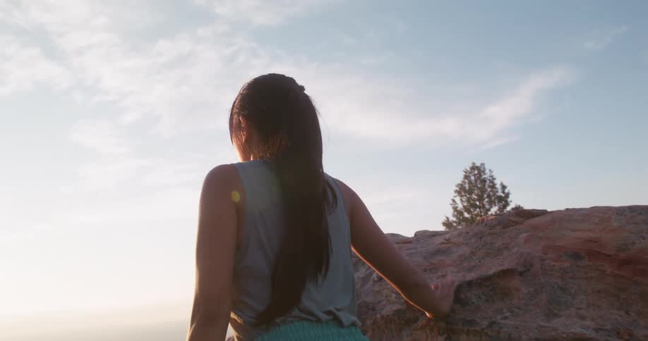 A woman with long dark hair is seen from behind climbing over a large rock formation under a bright sky with a sunset glow