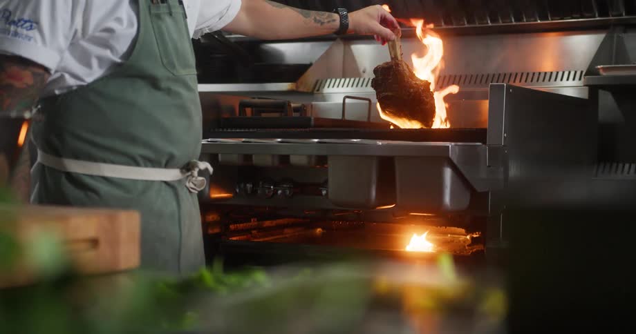 A chef cooks meat over open flames on a commercial stove in a restaurant kitchen