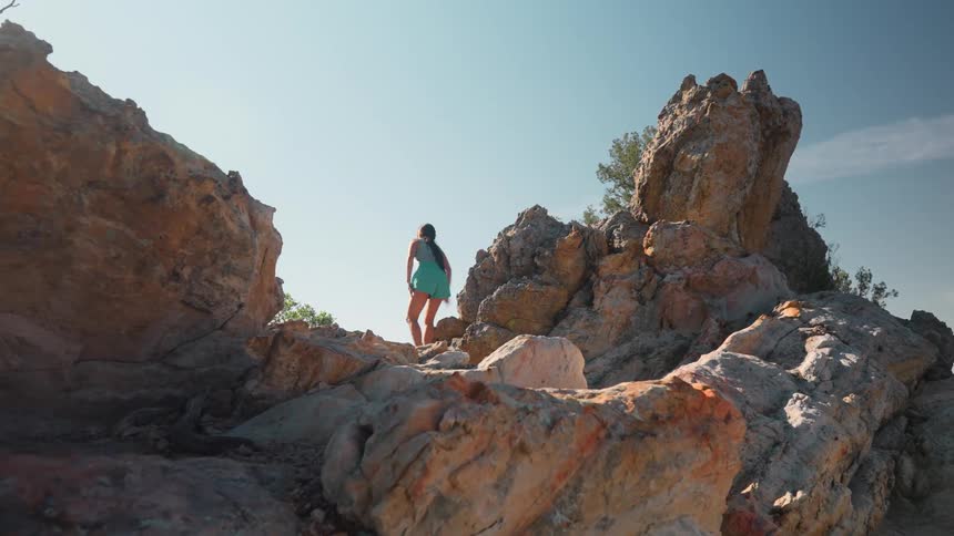 A woman wearing a teal outfit hikes among large rugged rock formations under a clear blue sky surrounded by scattered vegetation