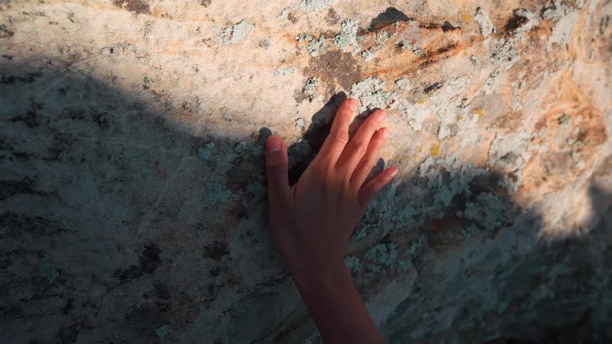 A persons hand is gently touching a textured rock surface sunlight and shadows play across the rock highlighting its uneven surface and the subtle movements of the hand