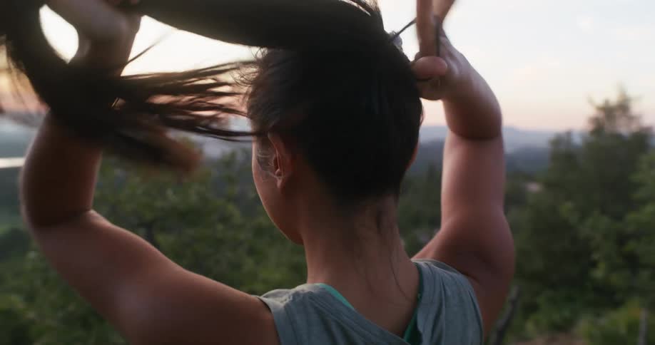 A young woman ties her hair while standing on top of a cliff at midday