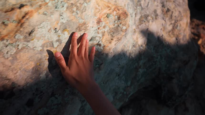 A persons hand rubs against a rock inside a cave with sunlight partially reflecting on the rock