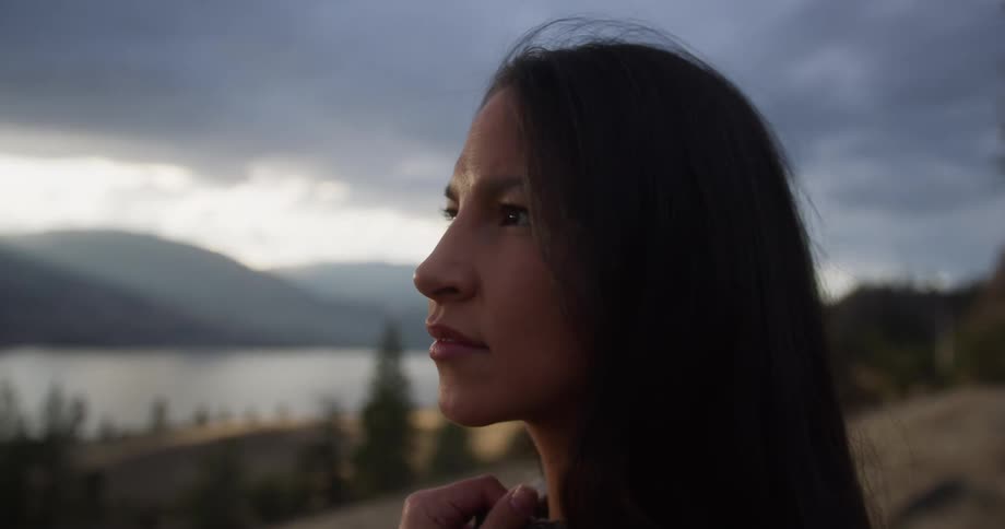 A woman gazes thoughtfully into the distance with a serene mountain lake and cloudy sky behind her