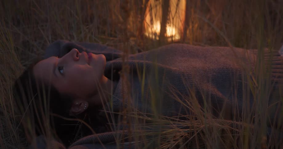 A woman lies in tall grass at dusk gazing upward near a glowing lantern