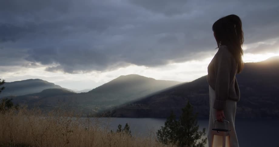 A woman stands overlooking a lake and mountains during sunset