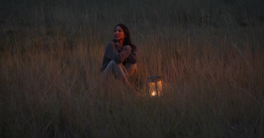 A woman sits in the middle of a field with a lantern and very tall grass