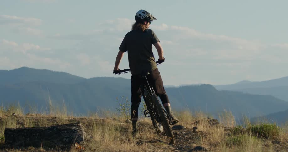 A mountain biker stands on a scenic trail overlooking distant blue mountains