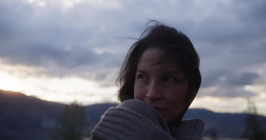 A woman stands peacefully with her eyes closed enjoying the breeze as the sky glows softly at dusk