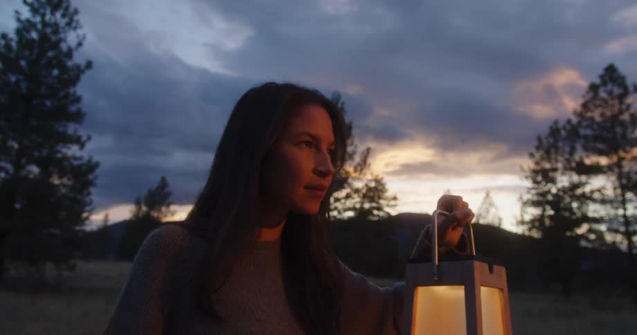 A woman holds a glowing lantern as evening falls over a forested landscape