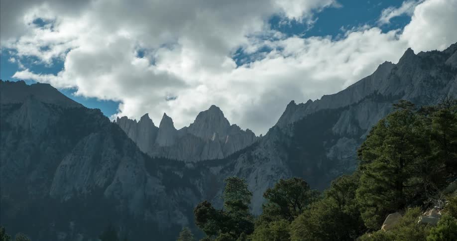 A dramatic timelapse view of Mount Whitney rises under a cloudy sky with trees in the foreground