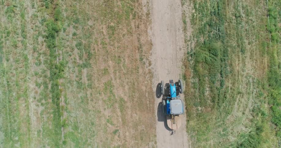 An aerial topdown view shows a blue tractor driving along a dirt path between two green crop fields