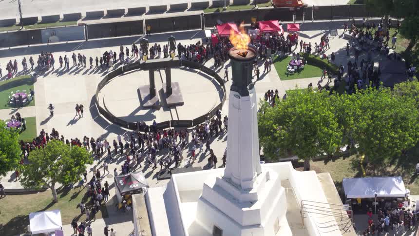 The Los Angeles Memorial Coliseum with its iconic olympic cauldron lit hosts a large crowd walking to and fro during a significant event or commemoration at midday