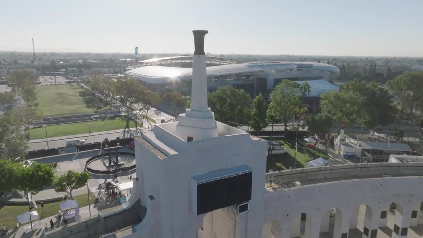 An aerial view shows the entrance and Olympic torch tower of the Los Angeles Memorial Coliseum with USC and fight on banners visible surrounded by trees seating and people