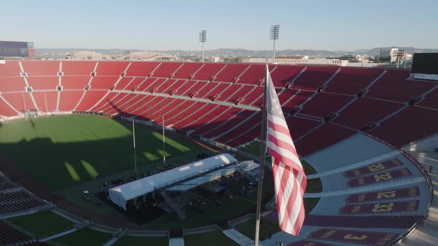 An empty Los Angeles Memorial Coliseum with red seating is shown under clear skies with an American flag prominently waving in the foreground