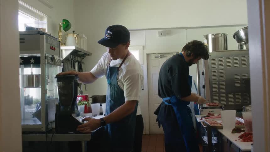 Two men work in a kitchen one blending and one preparing food