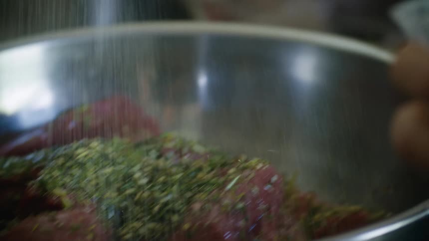 A hand is sprinkling salt over raw meat covered in herbs in a metal bowl