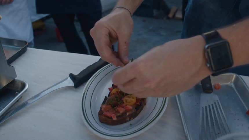 A chef garnishes a piece of bruschetta on a plate at a prep station