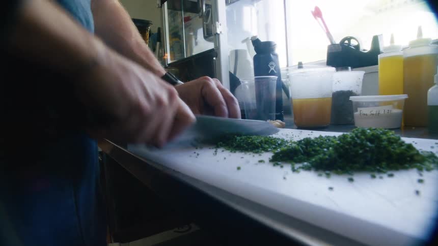 A person is chopping fresh herbs on a cutting board in a welllit kitchen with containers of ingredients and utensils visible