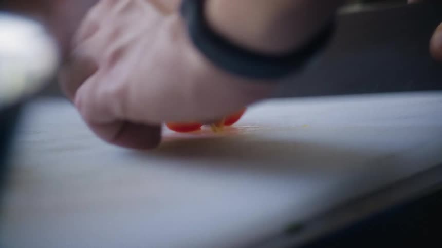 A closeup of hands chopping small red cherry tomatoes on a white cutting board a knife is visible on the right and the scene implies food preparation possibly in a kitchen setting