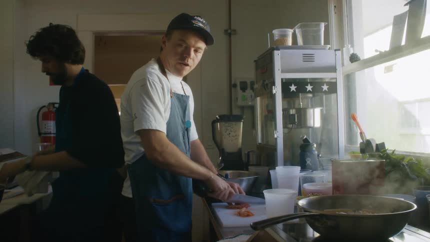 Two chefs work together in an indoor kitchen preparing food beside a steaming pan