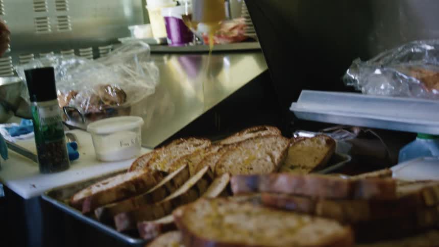 From a low angle on a kitchen prep line a chef drizzles sauce from a squeeze bottle while a tray of toasted bread sits in the foreground