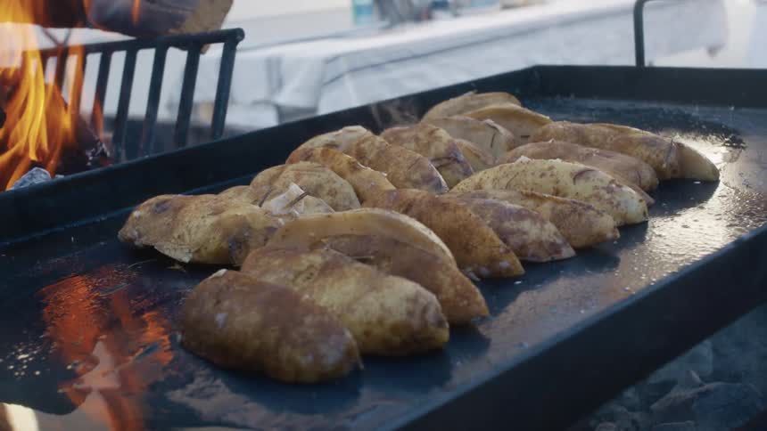 Sliced potatoes are grilling on a flattop grill beside an open flame in an outdoor cooking setup
