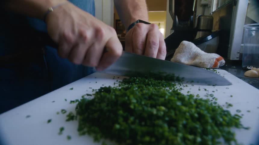 A person chops green herbs finely on a white cutting board in a kitchen