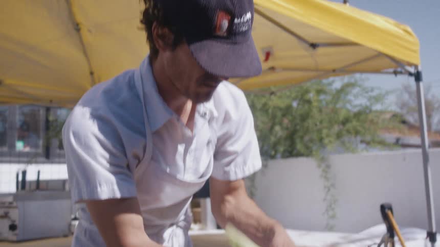 A chef wearing a hat and apron stands under an outdoor tent placing food on the grill at midday