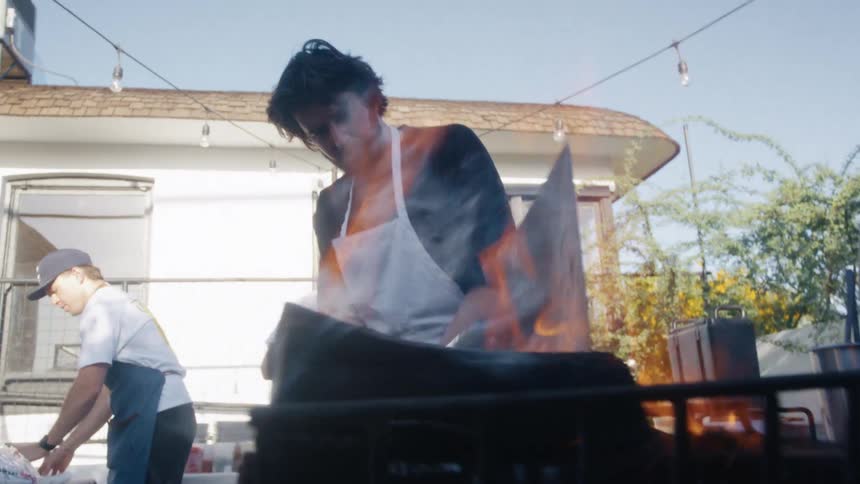 A man uses a shovel to break down large burning charcoal on a grill while two other men behind him prepare food items in the backyard of a building at midday