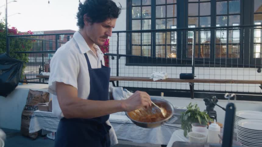 Man in apron holding frying pan with pasta in sauce in outdoor kitchen
