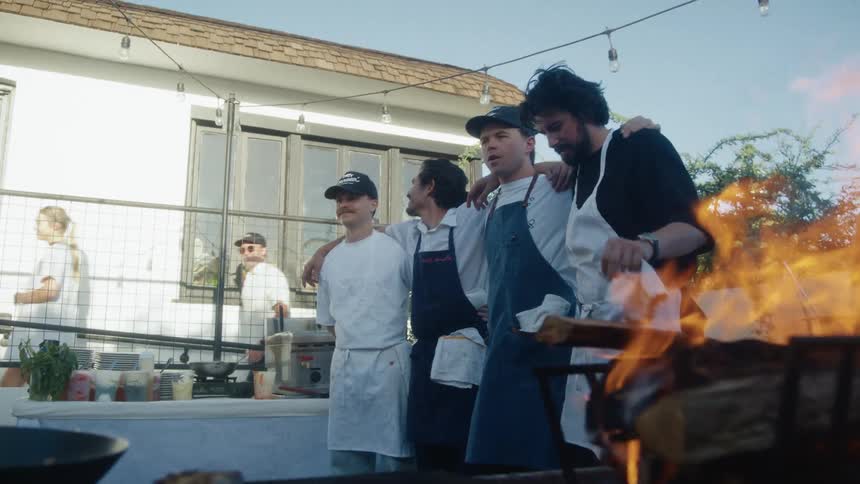 Four men hugging and wearing aprons inside an outdoor kitchen