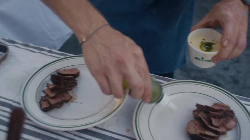 A person plates slices of grilled meat and drizzles sauce while holding a small cup