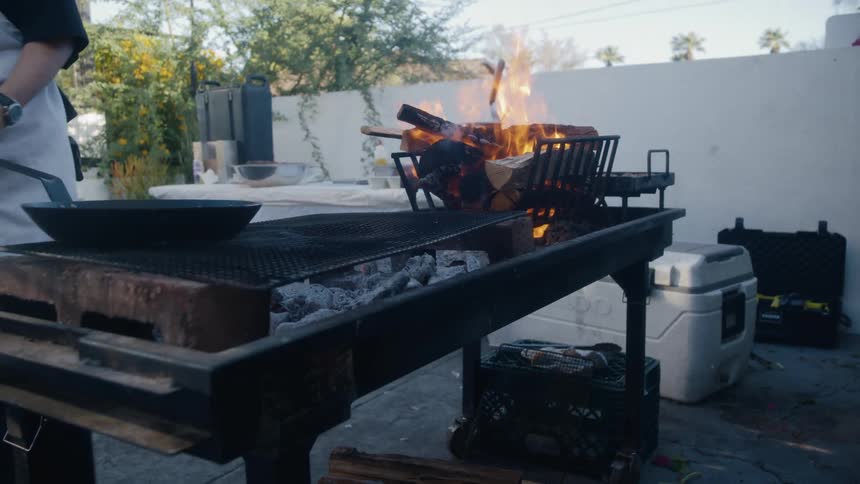 A person cooks over an outdoor woodfired grill where flames rise from a basket of burning logs and a skillet sits on the grate beside glowing coals