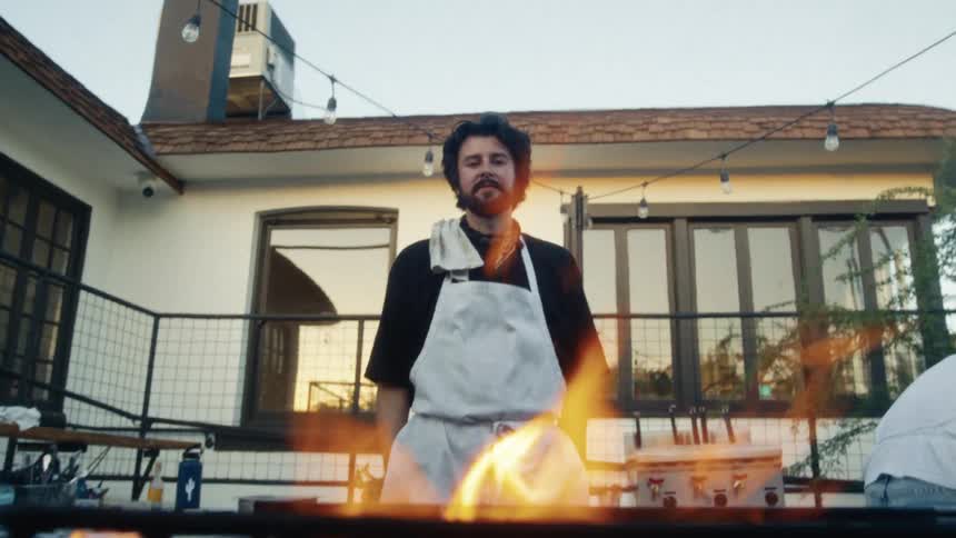 Bearded man wearing a white apron while standing in front of a lit barbecue