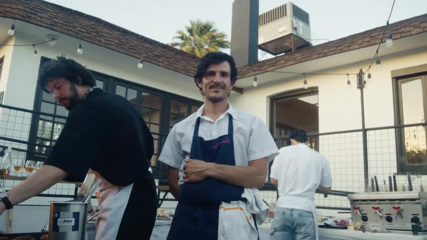 Chefs work at an outdoor kitchen setup while one smiles and poses for the camera