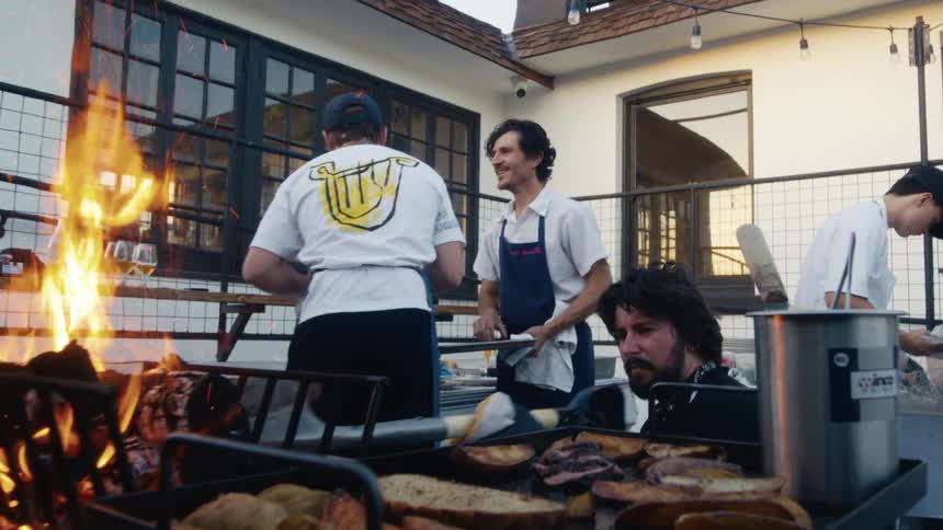 Chefs work together at an outdoor kitchen grilling food beside open flames