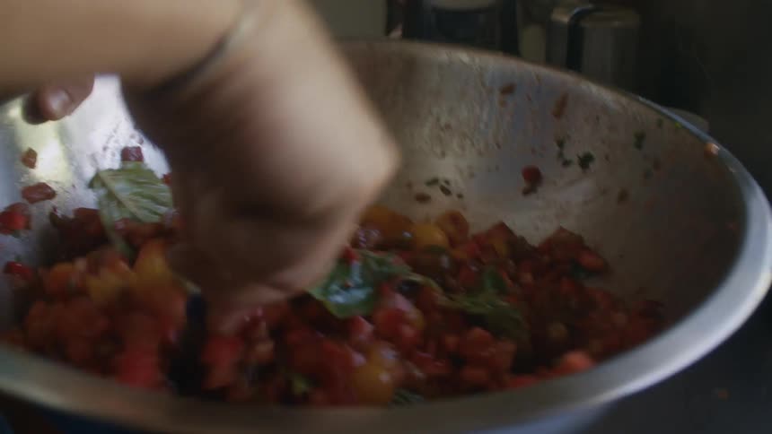 A hand stirs a vibrant mixture of diced vegetables in a large metal bowl