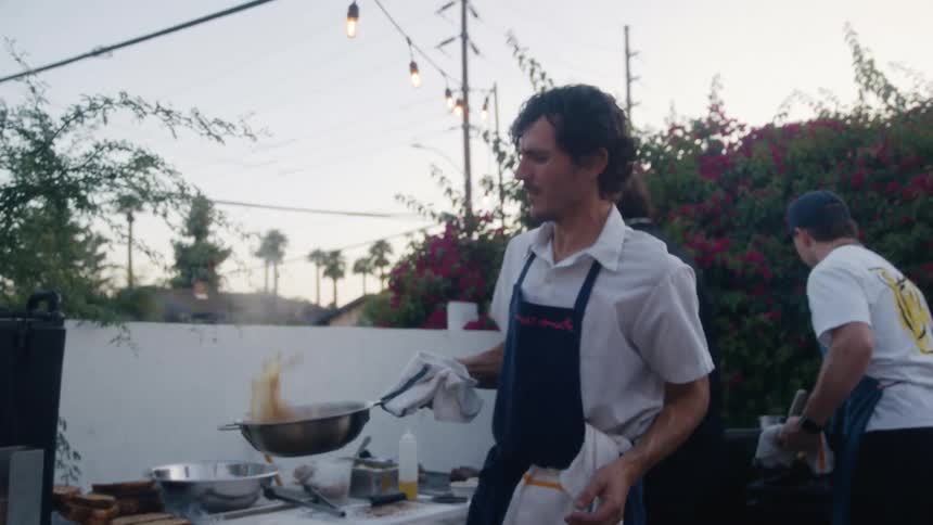 Three chefs cook outdoors at dusk with one flipping food in a pan