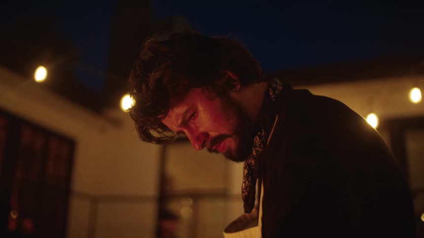 A man wearing an apron and neckerchief is focused on his task under warm string lights during the evening