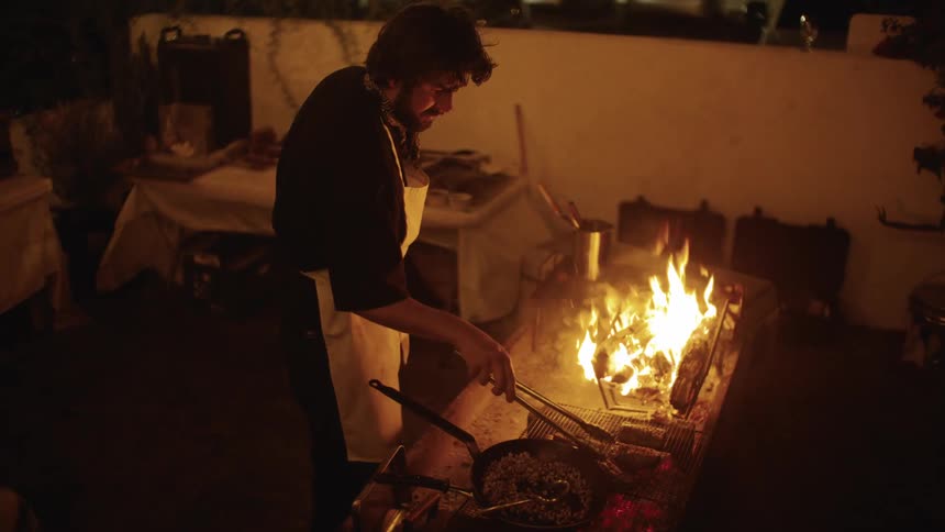 A man cooks over an open flame at an outdoor grill during nighttime