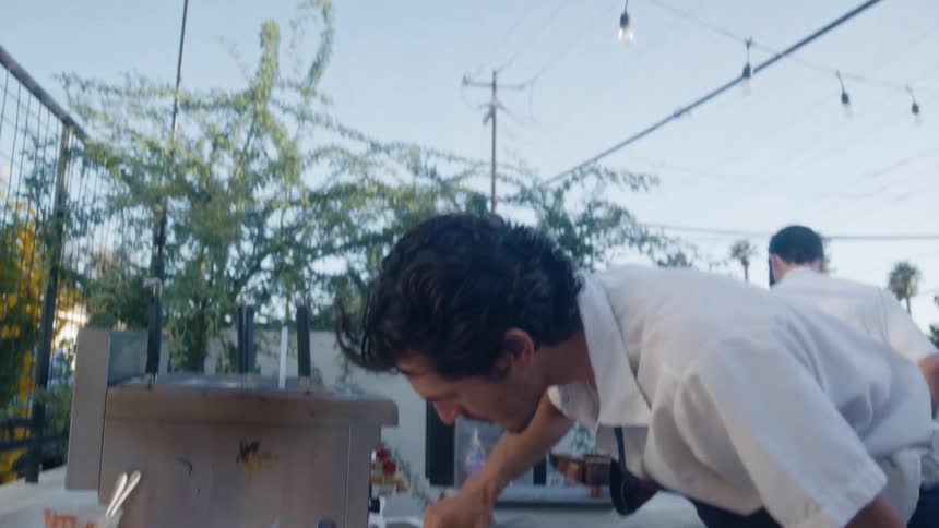A chef in an apron meticulously prepares food at an outdoor catering event adding sauces to bowls on a long table