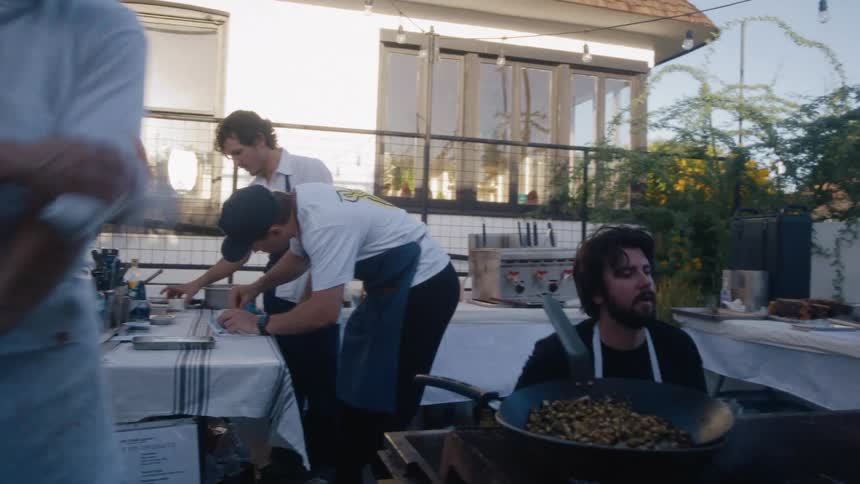 Four chefs work in an outdoor kitchen as one tends to a flaming grill in the foreground